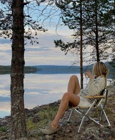 a woman sitting in a chair on the shore of a lake drinking from a cup