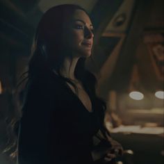 a woman with long dark hair standing in a dimly lit room looking off into the distance