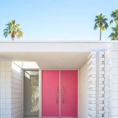 a pink door in front of a white building with palm trees behind it and the entrance