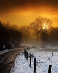 the sun is setting over a snowy road and fenced in area with snow on the ground