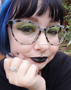 a woman with blue hair wearing glasses and piercings is posing for the camera in front of some bushes