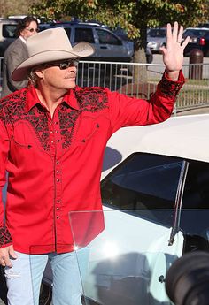 a man in a red shirt and cowboy hat standing next to a white car with his hand up