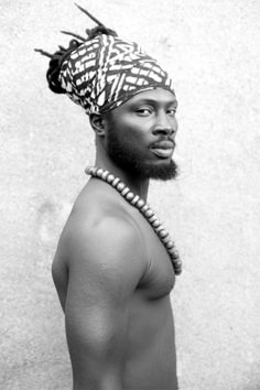 a man with a head scarf on and a necklace around his neck is standing in front of a concrete wall