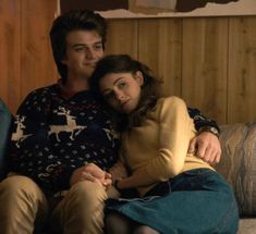 a young man and woman sitting on a couch next to each other in front of a wooden wall