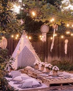 an outdoor dining area with lights strung over the table and pillows on the ground, in front of a wooden fence