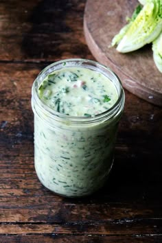 a glass jar filled with green sauce on top of a wooden table next to lettuce