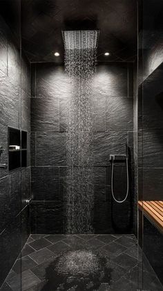 a shower head in the middle of a black tiled bathroom with wooden bench under it