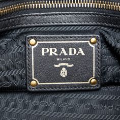 This Prada TessuTo handbag is made of black nylon canvas and has rolled leather indexes. The Bag is also decorated with gold -colored details. A golden Prada lettering shines on the front. The Bag Has a magnetic closure at the upper opening. Inside the Bag there are a zipper compartment and a slide compartment. The Bag can be in the hand or with a removable leather shoulder strap over the shoulder. Luxury Shoulder Bag With Logo Plaque And Top Handle, Luxury Shoulder Bag With Top Handle And Logo Plaque, Black Leather Bags With Logo Plaque, Luxury Shoulder Bag With Logo Plaque For Business, Gold Travel Bags With Logo Hardware, Luxury Tote Shoulder Bag With Logo Hardware, Gold Shoulder Bag With Logo Hardware For Travel, Gold Tote Bag With Logo Hardware, Luxury Business Bags With Gold-tone Logo Plaque