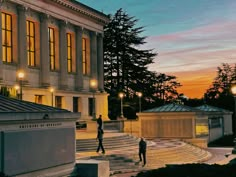 two people walking in front of a building at night with the sun setting behind them