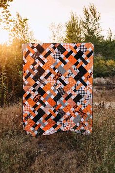 an orange and black quilt sitting on top of a grass covered field with trees in the background