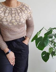 a woman standing in front of a potted plant wearing a sweater and black pants