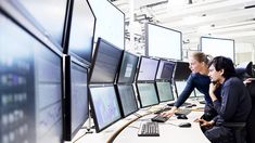 two women sitting in front of multiple computer screens on a white table and one woman is looking at the screen