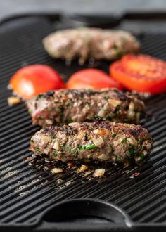 two hamburger patties are on the grill with tomatoes and other food items around them