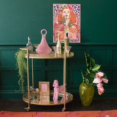 a gold shelf with pink flowers and other items on it next to a green wall