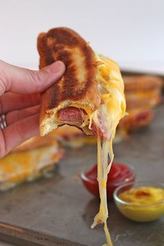 a person holding up a piece of pizza with cheese and ham on it, while dipping sauce in the background