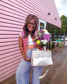 a woman standing on a wooden deck holding a white purse and flowers in her hand