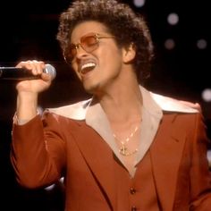 a man with glasses singing into a microphone and wearing a brown suit, white shirt and gold necklace