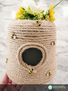 a hand holding a rope wrapped jar with bees and flowers in it on the table