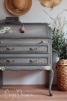 an old dresser painted in gray with flowers and leaves on the top, next to two hats hanging on the wall