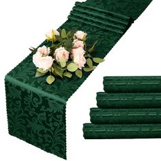 green table runner with roses and leaves on the top, next to four rolls of dark green satin