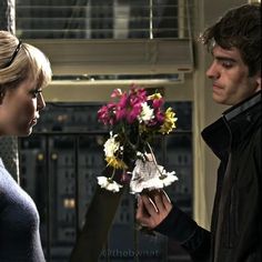 a man and woman standing next to each other in front of a window holding flowers