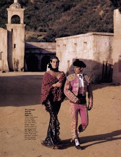 a man and woman dressed in costume walking down a dirt road next to an old building