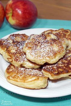 pancakes with powdered sugar are on a white plate next to two pieces of fruit