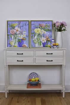 a white table with flowers and fruit on it in front of a framed art piece