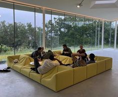 four people sitting on a yellow couch in front of large glass windows with trees outside