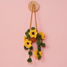 a crocheted flower arrangement hanging from a hook on a pink wall with green leaves and yellow flowers