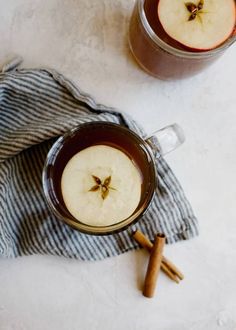 two glasses of apple cider with cinnamon sticks next to them on a white surface