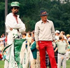 two men standing next to each other on top of a green golf ball field with people in the background