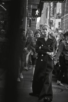 black and white photograph of woman in trench coat standing on sidewalk with crowd behind her