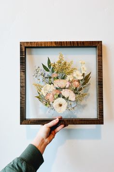 a person holding up a frame with flowers in it on the wall next to a white wall