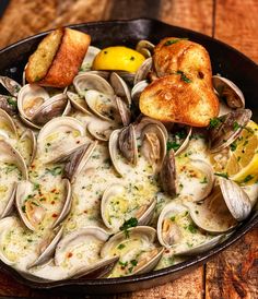 a skillet filled with clams and bread on top of a wooden table next to a lemon wedge