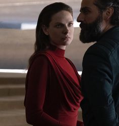 a woman in a red dress standing next to a man with long hair and beard