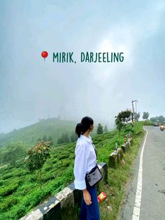 a woman standing on the side of a road next to a lush green hillside covered in fog