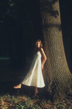 a woman in a white dress standing next to a tree with her feet on the ground