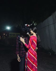 two people standing next to each other on a sidewalk at night with lights in the background