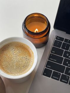 a cup of coffee sitting next to a laptop on a white table with a candle