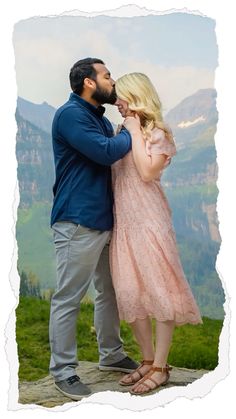 a man and woman standing next to each other on top of a hill with mountains in the background