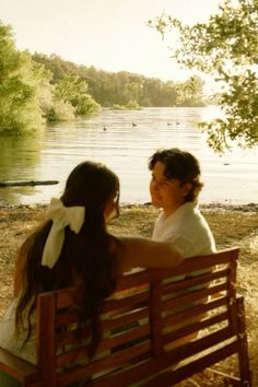 two people sitting on a bench next to a body of water with trees in the background