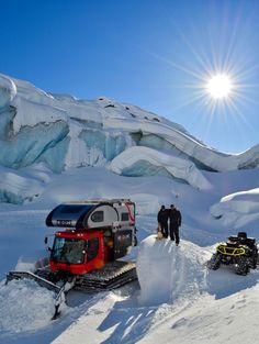 two people are standing in the snow next to some vehicles and an iceplow