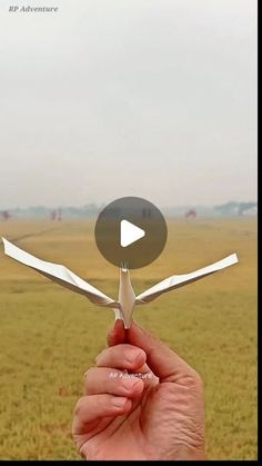 a hand holding scissors in the middle of a field with an airplane flying above it