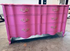 a pink dresser with gold handles in a warehouse or office space, it is painted bright pink and has ornate details on the drawers