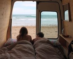 two people laying in bed looking out an open window at the beach and ocean from inside a camper van