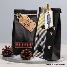 two black paper bags with gold and white designs on them, one has a pine cone next to it