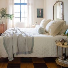 a bed with white linens and pillows in a bedroom next to a dresser, mirror and potted plant