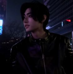 a young man standing in front of a tall building at night wearing a leather jacket