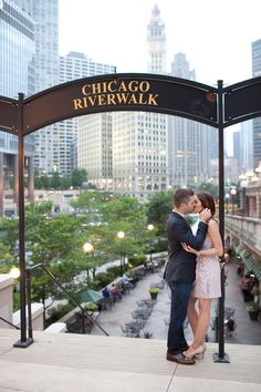 a man and woman standing under a chicago riverwalk sign with the city in the background